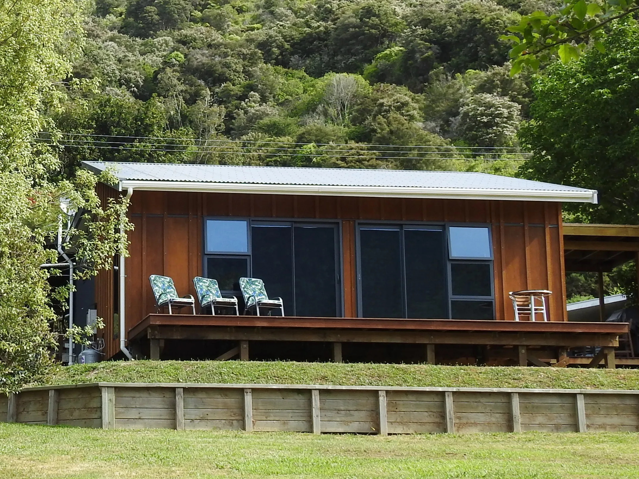 The hand-built timber cabin front-on — full-height glass and a timber deck with chairs, set against the native bush