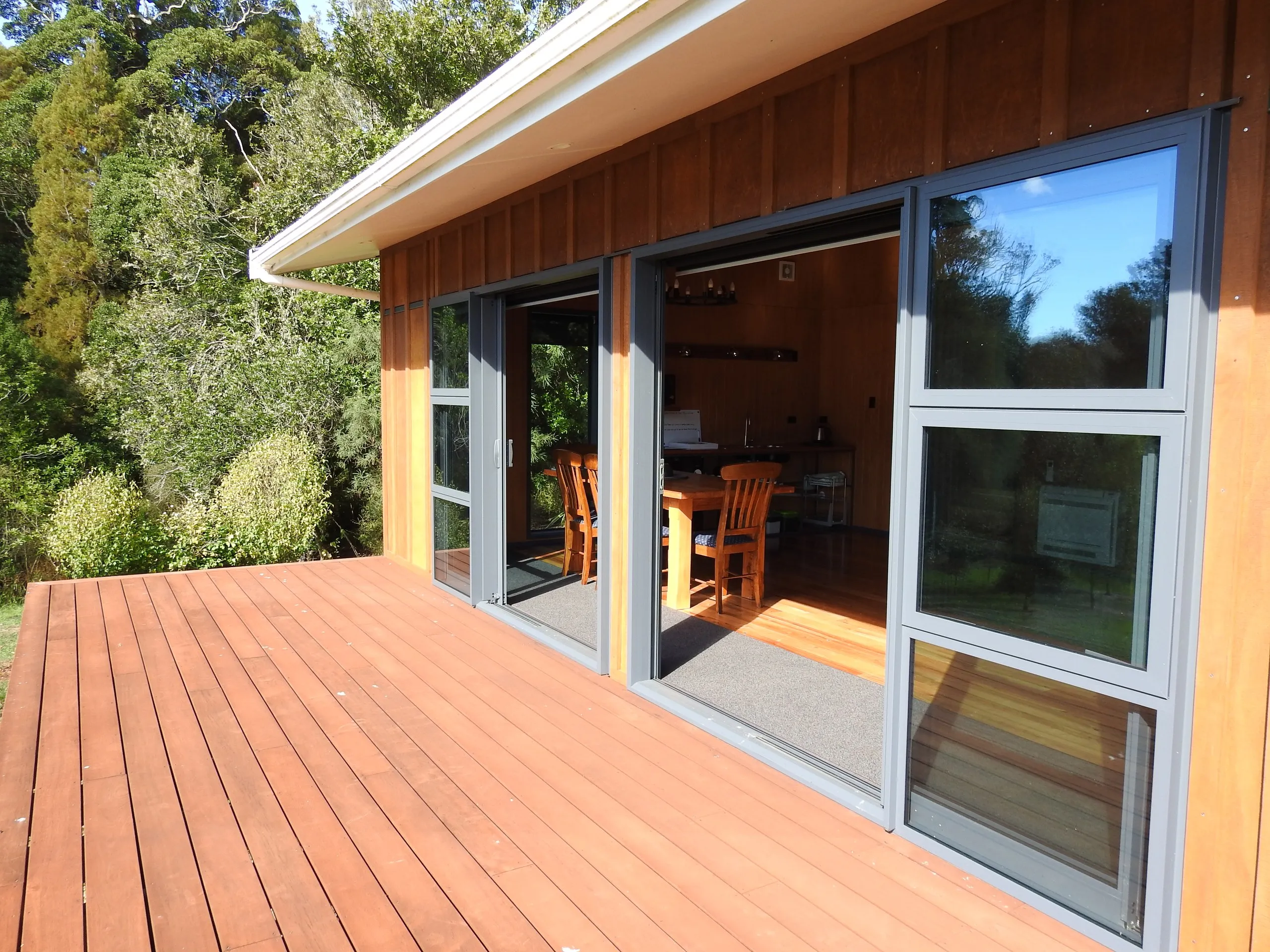 The cabin’s timber front deck, sliding glass doors open to the dining room, framed by native bush